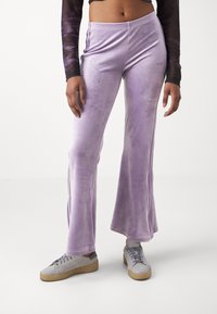 Kvinna iklädd lila velour byxor med vida ben, grå sneakers och en långärmad cropped topp, stående mot en neutral vit bakgrund.