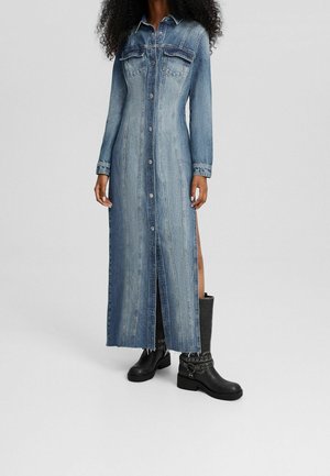 Kobieta w długiej, zapinanej na guziki sukience z denimu z kieszeniami na piersi, bocznymi rozcięciami oraz czarnych, sięgających połowy łydki botkach z paskami i srebrnymi przelotkami.