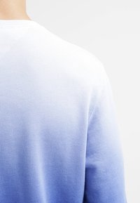 Acercamiento a la parte superior de la espalda de una persona que lleva un suéter de gradiente blanco a azul claro sobre un fondo ligero y liso.