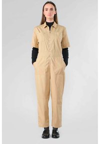 Combinaison beige à manches courtes, avec fermeture éclair à l'avant, deux poches poitrine et poches latérales. Tissu texturé et coupe décontractée. T-shirt noir visible en dessous.