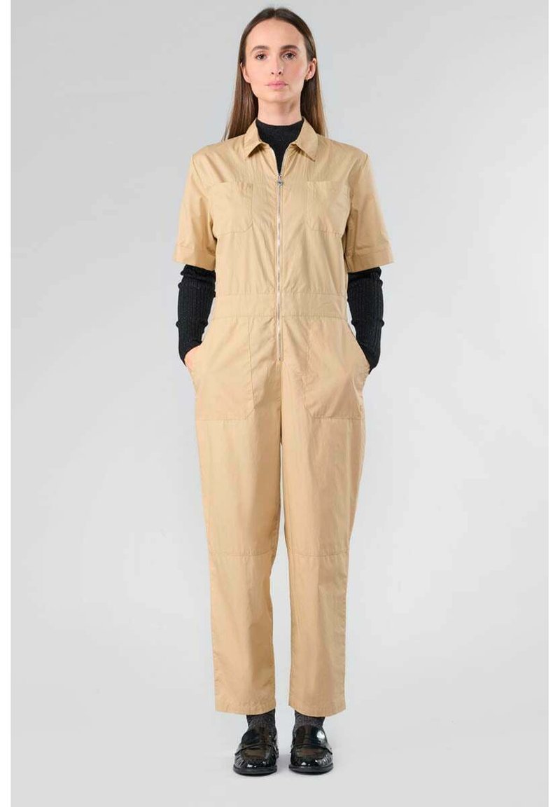 Combinaison beige à manches courtes, avec fermeture éclair à l'avant, deux poches poitrine et poches latérales. Tissu texturé et coupe décontractée. T-shirt noir visible en dessous.