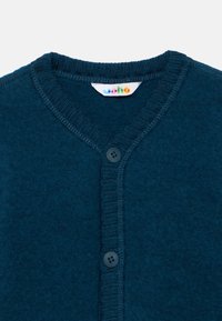 Námořnicky modrý cardigan s výstřihem do V, vyrobený z měkkého materiálu, s žebrovaným límcem a zapínáním na knoflíky. Obsahuje barevnou značkovou etiketu.