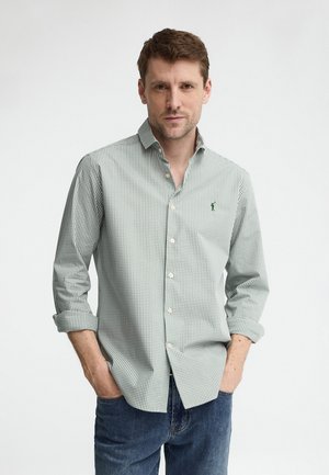 Hombre con camisa de cuadros verde claro y blanca abotonada con las mangas remangadas y vaqueros azules, manos en los bolsillos, fondo neutral.