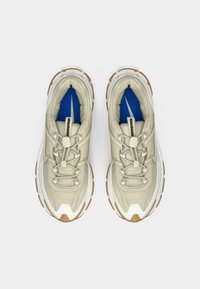 Um par de ténis beiges e brancos com solas castanhas grossas, atacadores elásticos sem nó, e palmilhas azuis com o logótipo branco visível dentro.