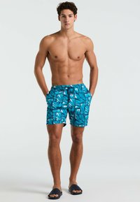 Shorts de bain pour hommes avec un motif tropical de feuilles bleu et vert vif, dotés d'une taille élastique, de poches latérales et d'une coupe décontractée.