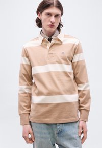 Νέος άνδρας με μακριά μαλλιά, ντυμένος με μακρυμάνικο πόλο πουκάμισο μπεζ με λευκές ρίγες και ανοιχτό μπλε τζιν, στέκεται μπροστά σε απλό φόντο.