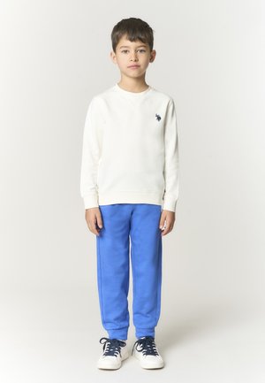 Ragazzo in piedi, indossa una felpa bianca a manica lunga con piccolo logo, pantaloni blu e scarpe da ginnastica bianche con lacci scuri, su sfondo chiaro uniforme.