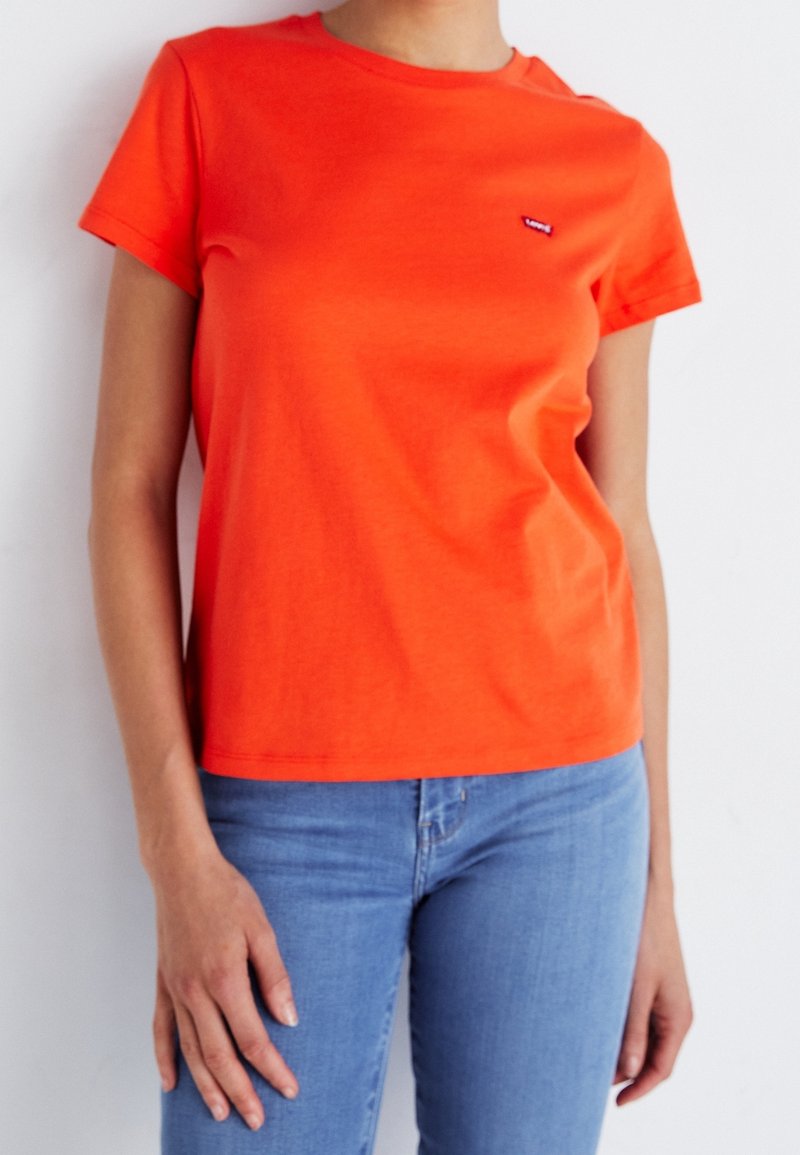 Personne portant un t-shirt orange vif à manches courtes avec un petit logo sur la poitrine et un jean bleu clair sur un fond uni.