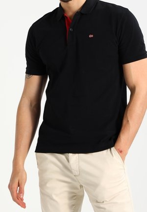 Polo noir avec un col à boutons, présentant un accent rouge sous le col et un petit logo sur la poitrine. Porté avec un pantalon beige.