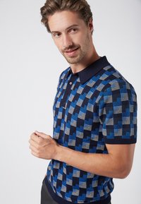 Tummansininen ja sininen lyhythihainen polo-paita, jossa on geometrinen kuvio, varustettuna kauluskannalla ja kolmella napilla. Sileä pinta.