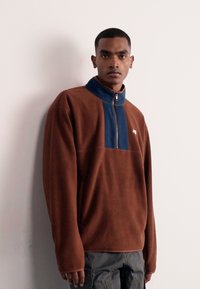 Pullover in pile marrone con collo a zip quarti blu navy, caratterizzato da un logo discreto sul petto e maniche dal taglio rilassato.