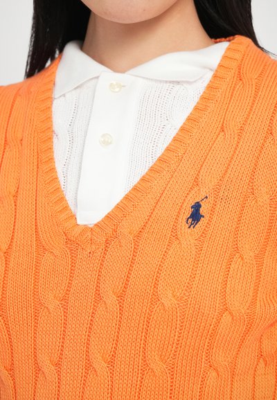 Oranža adīta veste ar V veida izgriezumu un kabeļu rakstu, valkāta virs baltas krekla ar apkakli. Uz krūtīm ir neliels zils logo.