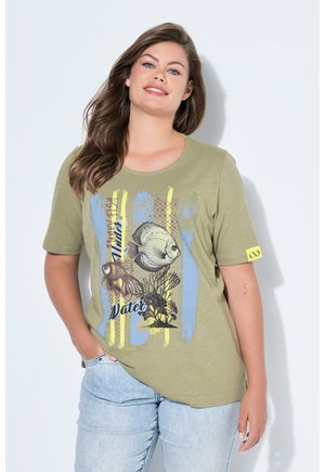 Lächelnde Frau mit langen braunen Haaren, die ein olivgrünes T-Shirt mit Grafik von Fischen und Wasserpflanzen sowie hellblaue Jeans trägt.