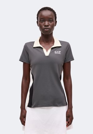 Femeie purtând un tricou polo gri cu mâneci scurte, guler alb și logo EA7 Emporio Armani, asortat cu o fustă albă, pe un fundal simplu.