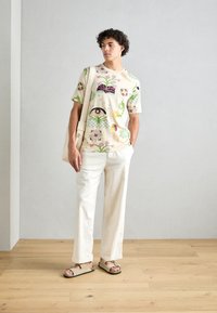 Short-sleeve cream shirt featuring colorful floral and geometric patterns, paired with light-colored, wide-leg trousers and flat sandals.