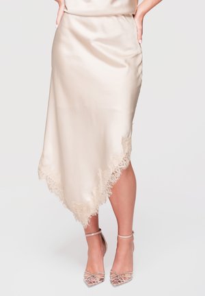 Femme portant une jupe en satin crème avec bordure en dentelle et des escarpins pointus avec brides à la cheville, debout les mains sur les hanches devant un fond uni.