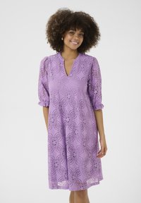 Lavandas krāsas mežģīņu kleita ar V veida izgriezumu un puffa piedurknēm, ar ziedu rakstiem un līdz ceļiem garuma, izceļot smalkas detaļas.