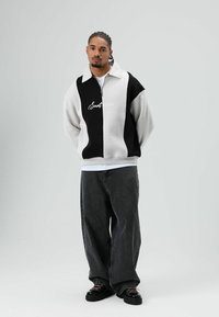 Wit-zwart sweatshirt met ritssluiting, een grote kraag en het geborduurde 'Savantessence' logo, gecombineerd met losse donkere spijkerbroeken en zwarte schoenen.