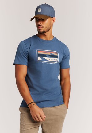 Blauw katoenen T-shirt met een grafisch ontwerp van golven en zonsondergangkleuren. Draagt beige gestructureerde broek en een bijpassende blauwe pet.