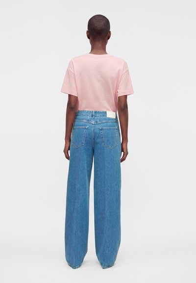 Personne debout de dos face à la caméra, portant un t-shirt rose pâle à manches courtes et un jean bleu taille haute à jambes larges, sur un fond uni.