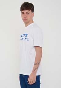 T-shirt bianco in cotone con testo e stampa grafica blu; colletto girocollo, maniche corte, taglio rilassato, indossata con jeans in denim blu.