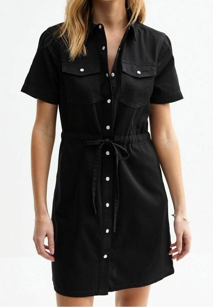 Femme portant une robe noire à manches courtes avec des poches poitrine et une ceinture nouée à la taille, debout devant un fond blanc uni.