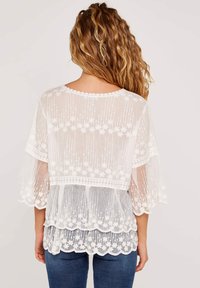 Vit genomskinlig blus med blommönster, vågig nederkant och vida ärmar. Har textil med struktur och intrikata detaljer på baksidan.