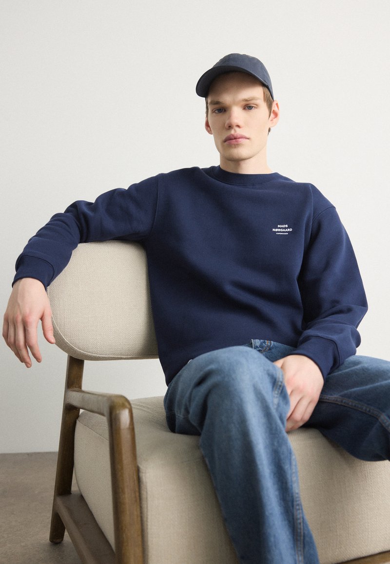 Σκούρος μπλε φούτερ με στρογγυλή λαιμόκοψη και μικρό λευκό λογότυπο. Κανονικά μπλε τζιν. Μοντέλο καθισμένο σε μια ανοιχτόχρωμη επενδεδυμένη καρέκλα.