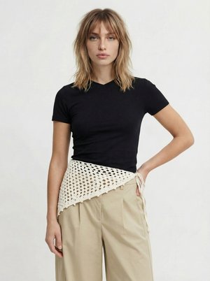 Woman with shoulder-length blonde hair wears a black short-sleeve top, beige wide-leg pants, and a white crocheted wrap tied at the waist.