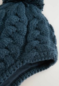 Mörkblå stickad mössa med ett texturerat kabelmönster. Har en mjuk, fluffig yta och en vikbar kant, prydd med en pom-pom på toppen.