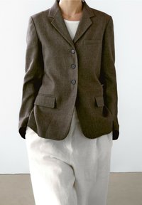 Ruskea räätälöity bleiseri, jossa on viisto kaulus, kolme nappia ja sivutaskut, yhdistettynä valkoisiin pellavahousuihin. Sujuva pinta ja moderni istuvuus.