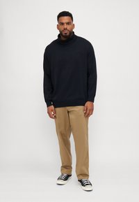 Schwarzer Rollkragenpullover aus weichem Stoff, mit lässigem Schnitt und langen Ärmeln, kombiniert mit beigen Hosen und schwarzen Sneakers.