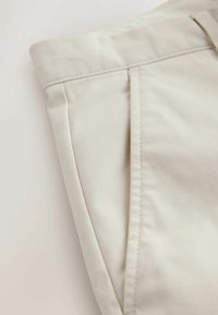 Pantalon beige sur mesure montrant une poche latérale, une ceinture et des coutures sur un fond clair.