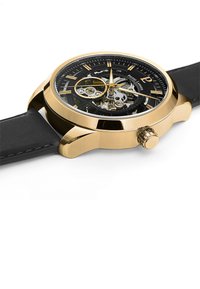 Montre en acier doré avec un bracelet en cuir noir. Le cadran squeletté révèle le mouvement, avec des accents dorés et argentés. Boîtier rond, couronne inclinée.