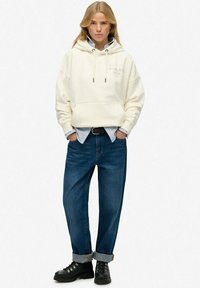 Haut à capuche couleur crème avec lacets, superposé sur une chemise rayée bleue et blanche, associé à un jean bleu ample et des bottes noires à lacets.