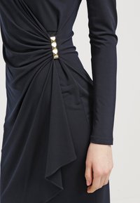 Robe noire à manches longues avec un détail froncé sur le côté, rassemblé par des clous dorés en forme de pyramide, dévoilant la taille et le bras droit.