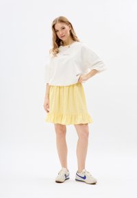 Witte oversized shirt met roze tekst, gecombineerd met een gele gerimpelde rok en witte schoenen met blauwe accenten. Eenvoudig, casual ontwerp.