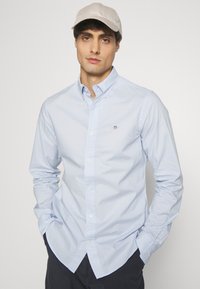  Lichtblauw overhemd met lange mouwen en knopen, voorzien van een klassieke kraag, een enkel borstlogo en witte knopen. Draagt een beige pet en donkere broek.