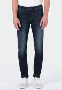 Homme portant un jean skinny bleu marine retroussé et des baskets blanches, se tenant devant un fond clair unicolore.