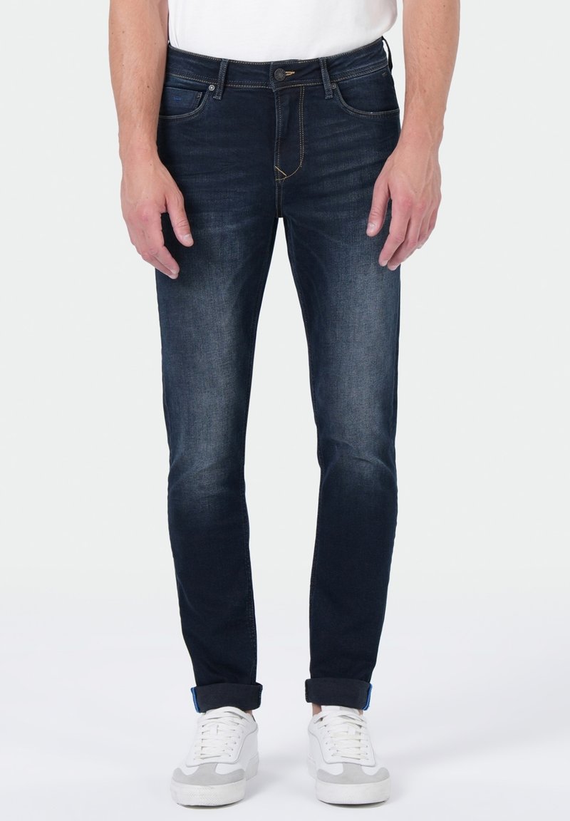 Homme portant un jean skinny bleu marine retroussé et des baskets blanches, se tenant devant un fond clair unicolore.