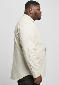 Beige langsliepend shirt met een mandarin kraag, met een rechte snit en subtiele textuur. Van opzij weergegeven.