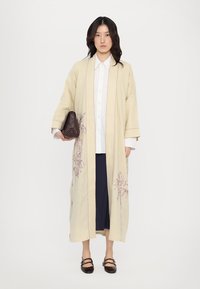 Krmovo sfarbený dlhý kimono s ružovými vyšívanými detailmi, širokými rukávmi a voľným strihom, v kombinácii s tmavými sukňami a hnedou kabelkou.