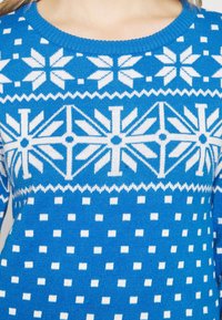 Sininen neulotutakki, jossa on valkoisia geometrisia kuvioita, mukaan lukien lumihiutaleet ja neliöt, ribbireunalla varustettu pyöreä kaula-aukko ja tekstuurinen kangas.