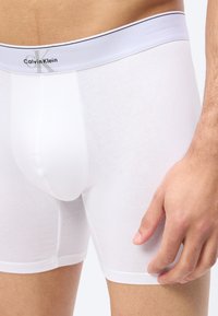 Άσπρα ανδρικά μποξεράκια Calvin Klein σε χρήση, εμφανίζοντας το λογότυπο στη μέση και κοντινή εικόνα του δεξιού χεριού που ακουμπάει στο μηρό.