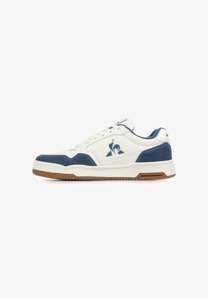 Zapatilla blanca de caña baja con detalles en ante azul, suela de goma marrón y un logo azul de un gallo en el lateral, mostrada desde el lado exterior.
