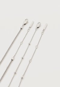 Τέσσερις ασημένιες αλυσίδες με κούμπωμα αστακού και μικρές ορθογώνιες πλακέτες, τοποθετημένες σε λευκό φόντο, με ποικίλα σχέδια συνδέσμων και λεπτομέρειες με χάντρες.