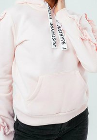 Sudadera con capucha de color rosa claro con mangas con volantes, bolsillo delantero y cordones negros y blancos con el texto "JUSTHYPE". Material de algodón suave.
