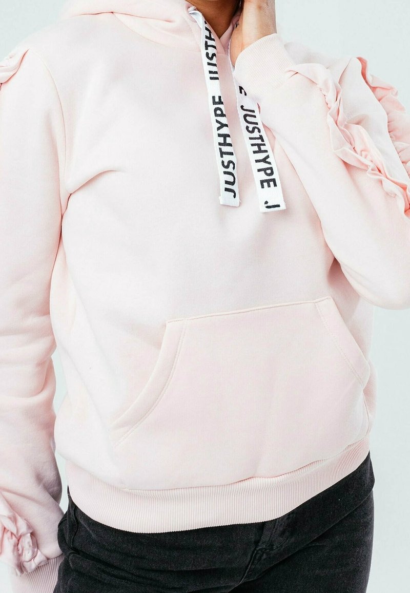 Sudadera con capucha de color rosa claro con mangas con volantes, bolsillo delantero y cordones negros y blancos con el texto "JUSTHYPE". Material de algodón suave.