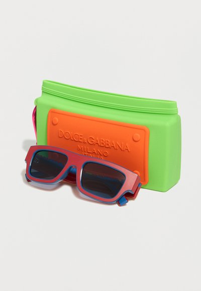 Μπλε και κόκκινα ορθογώνια γυαλιά ηλίου τοποθετημένα μπροστά από μια πράσινη και πορτοκαλί θήκη με το λογότυπο Dolce & Gabbana Milano.