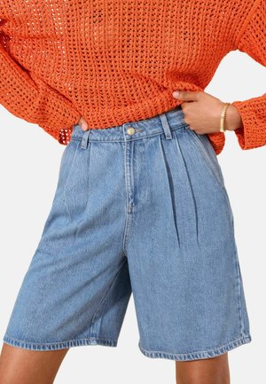 Kvinde iført lyseblå højtaljede, plisserede cowboyshorts og en orange strikket sweater med et guldarmbånd på højre håndled.